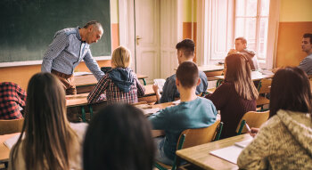 Blick von hinten in ein Klassenzimmer mit sitzenden Schülerinnen und Schülern. Vorne steht ein Mann.