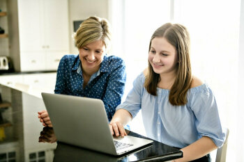 Eine junge und eine ältere Dame sitzen am Tisch vor einem Laptop. Sie lächeln.