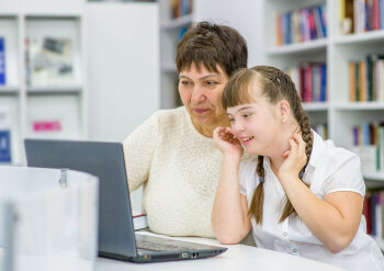 Ein Mädchen mit Down-Syndrom sitzt zusammen mit einer älteren Frau lächelnd vor einem Laptop.