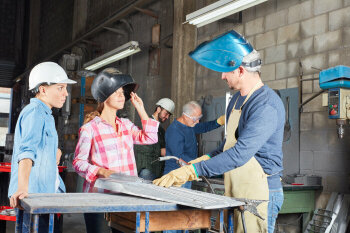 Zwei junge Frauen mit Helm und Schweißhelm reden mit einem älteren Mann, der ebenfalls einen Schweißhelm trägt.