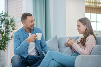 Ein älterer Mann und ein junges Mädchen sitzen zusammen auf der Couch. Beide halten jeweils einen Becher in der Hand.