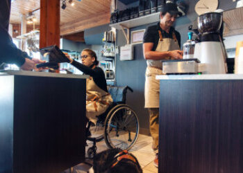 Eine Frau im Rollstuhl bedient einen Touchscreen in einem Geschäft. Daneben ein Mann an einer Kaffeemahlmaschine. 