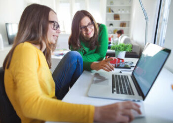 Ein Mädchen sitzt vor einem Laptop, eine Frau steht daneben und schaut mit auf den Bildschirm. 