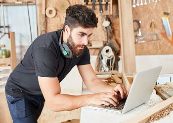 Auf einem Werkstatt-Tisch steht ein aufgeklappter Laptop an dem ein Mann in Arbeitskleidung arbeitet.