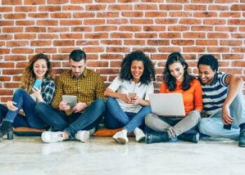 Drei Frauen und zwei Männer sitzen vor einer Mauer auf dem Boden und schauen auf zwei Handys, ein Tablet und einen Laptop.