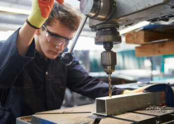 Ein junger Mann mit Handschuhen und Schutzbrille bedient eine industrielle Bohrmaschine. 
