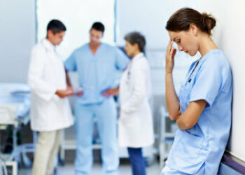 Eine Frau in Krankenhauskleidung steht nachdenklich an eine Wand gelehnt. Im Hintergrund reden drei Personen miteinander.