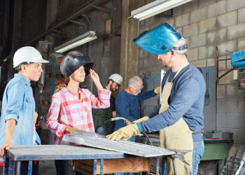 Zwei junge Frauen mit Helm und Schweißhelm reden mit einem älteren Mann, der ebenfalls einen Schweißhelm trägt.