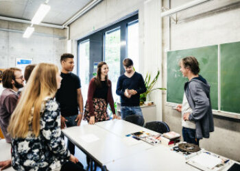 Mehrere Jugendliche stehen in einem Klassenzimmer um einen Tisch. Vor einer Tafel steht ein Mann. 