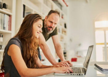 Eine Jugendliche sitzt lächelnd vor einem Laptop. Ein Mann schaut ihr dabei über die Schulter. 