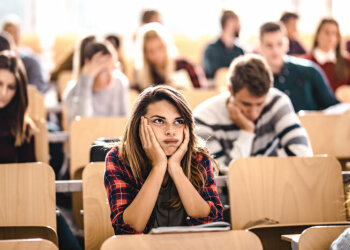 Ein Mädchen sitzt mit anderen Menschen in einem Vorlesungssaal und stützt den Kopf in die Hände. Dabei macht sie ein überfordertes Gesicht.