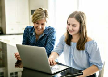 Eine junge und eine ältere Dame sitzen am Tisch vor einem Laptop. Sie lächeln.