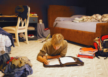 Ein Junge liegt in seinem Zimmer auf dem Boden und schreibt in einen Ordner. Um ihn herum liegen Bücher und Klamotten.