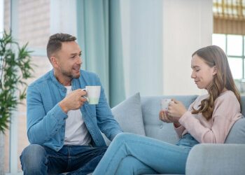 Ein älterer Mann und ein junges Mädchen sitzen zusammen auf der Couch. Beide halten jeweils einen Becher in der Hand.