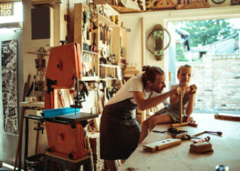 Ein Mann steht in einer Werkstatt und bearbeitet ein Holzstück. Auf dem Tisch sitzt ein Kind und fasst mit an. 