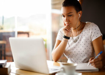 Eine junge Frau sitzt am Tisch, schaut auf den Laptop-Bildschirm vor ihr und macht sich Notizen. 