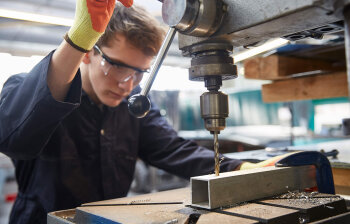 Ein junger Mann mit Handschuhen und Schutzbrille bedient eine industrielle Bohrmaschine. 
