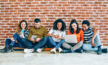 Drei Frauen und zwei Männer sitzen vor einer Mauer auf dem Boden und schauen auf zwei Handys, ein Tablet und einen Laptop.