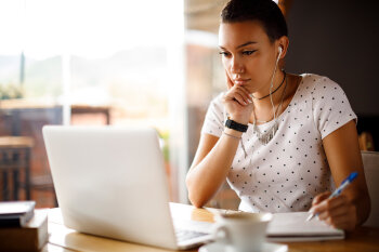 Eine junge Frau sitzt am Tisch, schaut auf den Laptop-Bildschirm vor ihr und macht sich Notizen. 