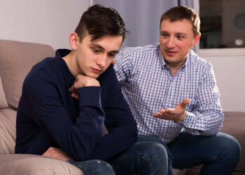 Ein Junge und ein Mann sitzen auf der Couch. Der Mann beschwichtigt den traurigen Jungen