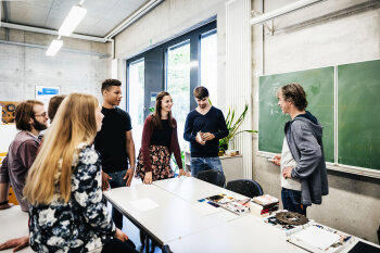 Mehrere Jugendliche stehen in einem Klassenzimmer um einen Tisch. Vor einer Tafel steht ein Mann. 