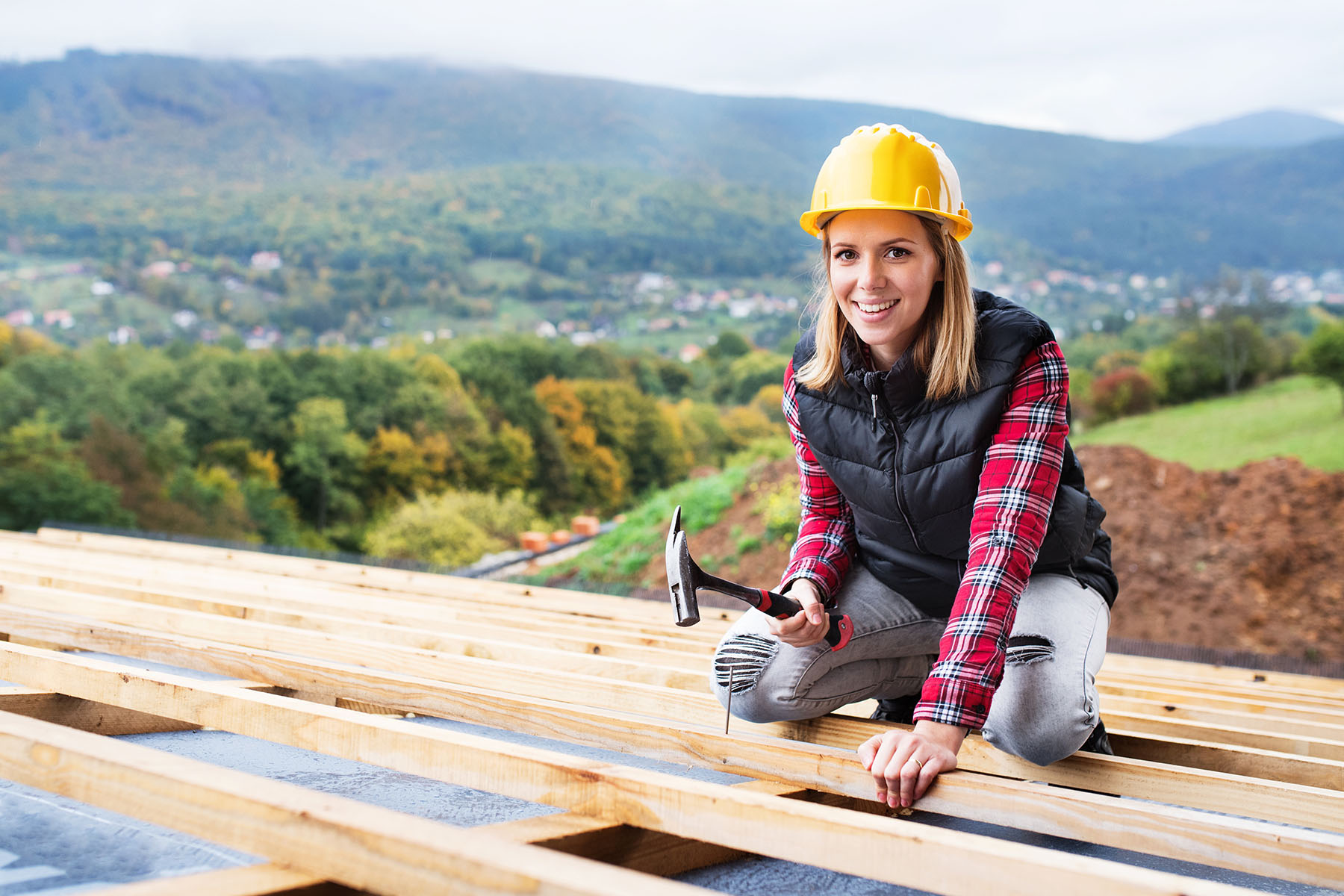 Eine junge Frau kniet auf einer Dachkonstruktion. In der rechten Hand hält sie einen Hammer.