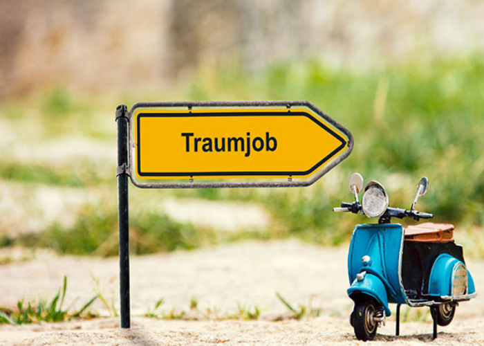 Auf einem gelben Wegweiser-Schild steht das Wort "Traumjob". Daneben steht ein kleiner Modell-Motorroller.