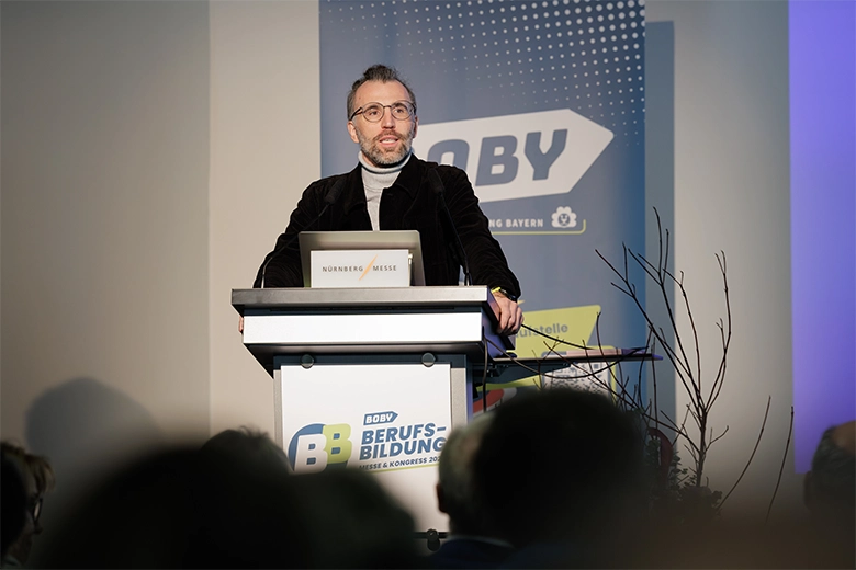 Ein man steht an einem Rednerpult und spricht zu Menschen, deren Köpfe nur von hinten zu sehen sind