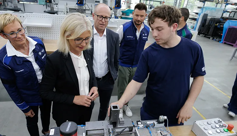 Mehrere Personen stehen in einer hellen Werkstatt um einen Arbeitstisch mit technischer Anlage; ein junger Mann im blauen Shirt demonstriert die Maschine. Blau, Grau und Weiß dominieren, die Szene wirkt sachlich und konzentriert.