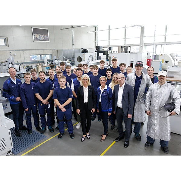 Große Gruppe von Mitarbeitenden in blauer Arbeitskleidung in einer hellen Industriehalle, vorne einige Personen in Anzug und Schutzmantel, sachliche Gruppenaufnahme.