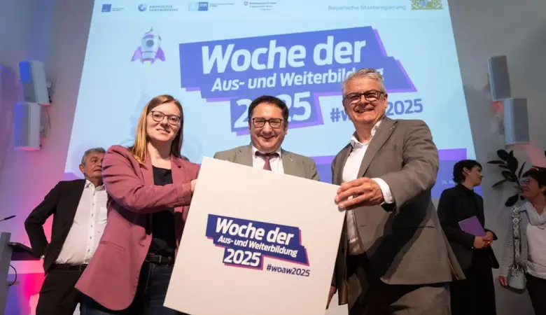 Bayerns Wirtschaftsstaatssekretär hält ein großes Bild mit dem Logo der Woche der Aus- und Weiterbildung.