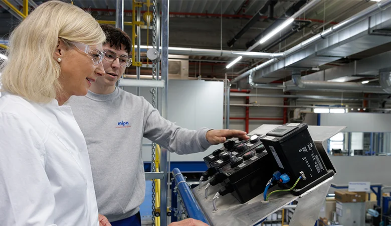 Ein Azubi und die Ministerin betrachten in einer hell beleuchteten Industriehalle ein schwarzes technisches Gerät auf einer Metallhalterung.