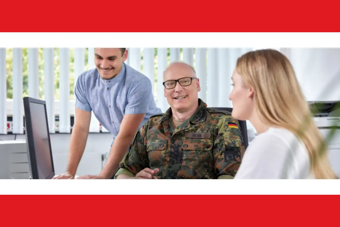 Ein  Mann in Soldatenuniform sitzt in der Mitte. Links von ihm steht ein Mann an einem Computer. Rechts eine junge Frau von der Seite.